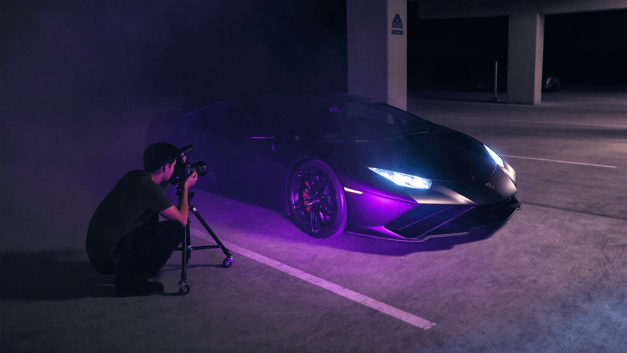 A photographer shooting a parked matte-black Lamborghini lit with purple rim light on a night set